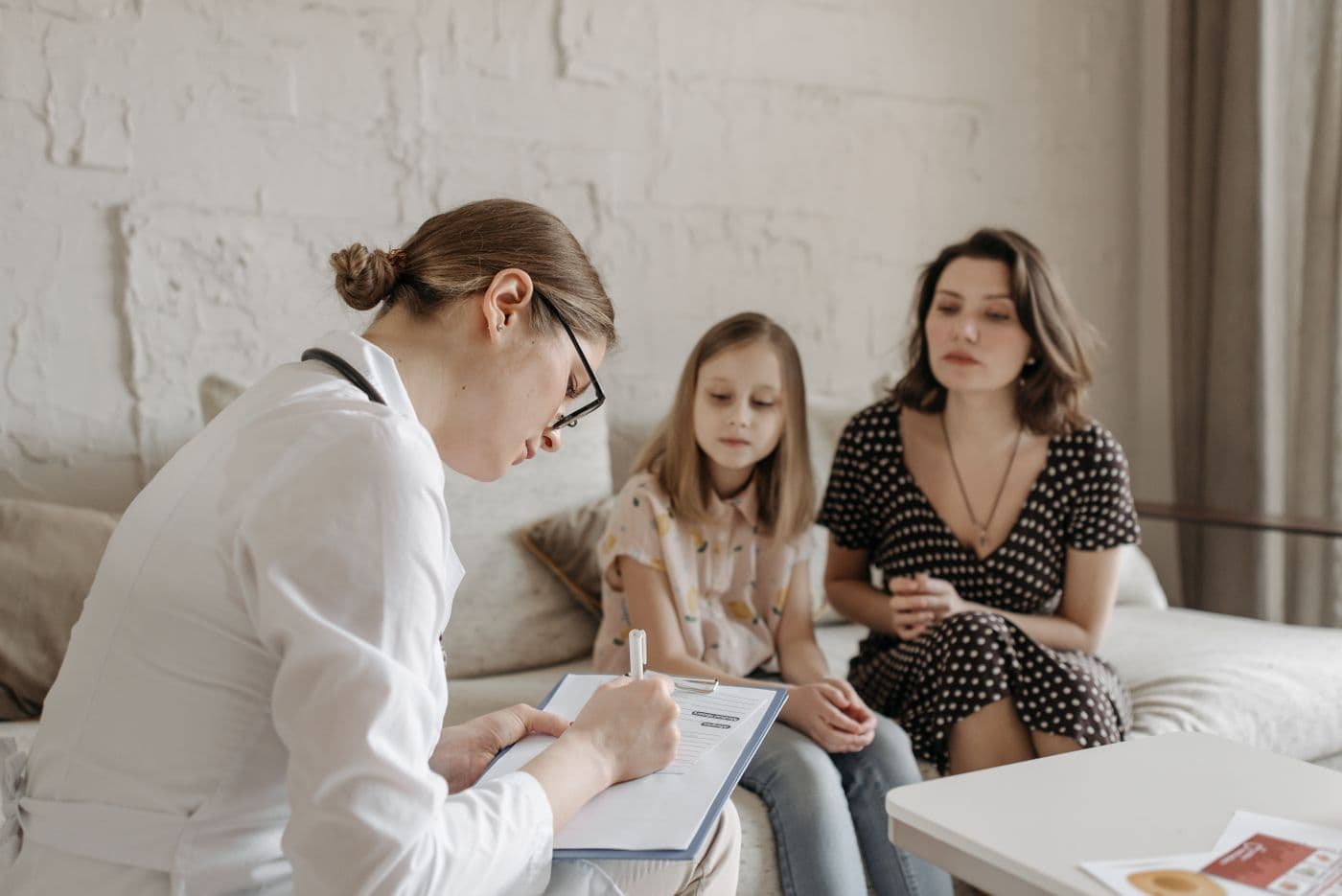 Médica conversa com uma mãe e sua filha durante uma consulta, explicando um documento em uma prancheta